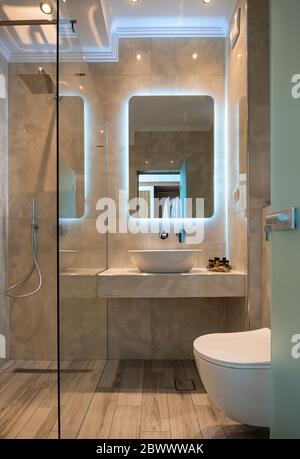 Vue sur la façade de la salle de bains moderne en marbre beige avec lavabo, miroir de lumière, cabine de douche en verre, toilettes murales, parquet Banque D'Images