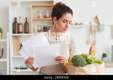 Jeune femme tenant des documents et vérifiant les produits dans un sac en papier en se tenant dans la cuisine Banque D'Images
