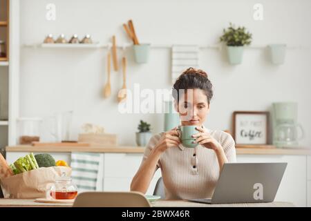 Une jeune femme buvant du café le matin et regardant l'écran d'un ordinateur portable tout en étant assise dans la cuisine à la maison Banque D'Images