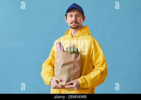 Portrait d'un jeune homme en uniforme tenant un sac en papier avec des produits sur fond bleu, il livrant de la nourriture Banque D'Images