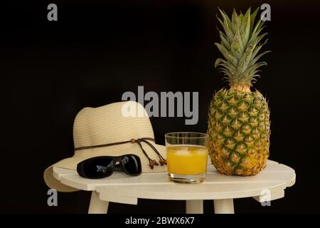 Ananas entier, jus de ananas, chapeau de soleil et lunettes de soleil sur table en bois blanc avec fond noir Banque D'Images