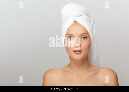 Portrait de jolie dame avec serviette blanche sur la tête sans maquillage avec des taches transparentes sous les yeux regardant rêveusement dans l'appareil photo sur fond gris Banque D'Images