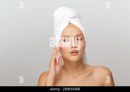 Portrait de jeune femme bouleversée avec une serviette blanche sur la tête sans maquillage avec des taches transparentes sous les yeux regardant soigneusement de côté sur le gris Banque D'Images