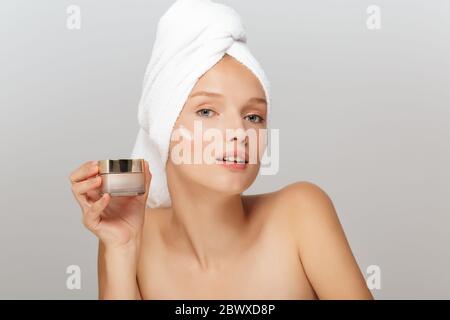 Portrait de jeune femme attrayante avec serviette blanche sur la tête sans maquillage tenant la crème du visage à la main tout en regardant avec soin dans l'appareil photo avec la crème Banque D'Images