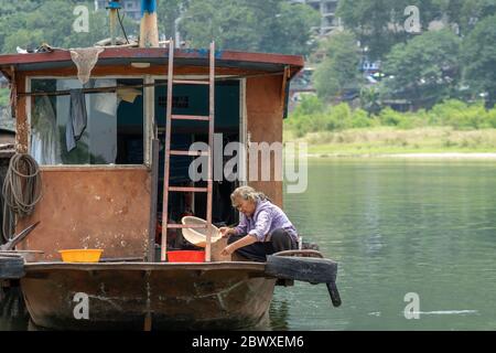 Yangshuo, Chine - août 2019 : femme chinoise vivant dans un bateau amarré sur le rivage de la rivière Li, utilisant un bol en plastique pour le lavage et la lessive Banque D'Images