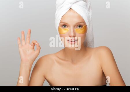 Portrait de jeune femme attrayante avec serviette blanche sur la tête sans maquillage avec taches jaunes sous les yeux, montrant le geste ok tout en regardant dans rêveur Banque D'Images