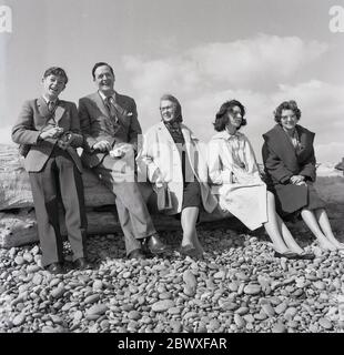 Années 1950, historique, photo de famille, l'homme portant un costume, son fils dans une veste et cravate et les femmes portant des manteaux assis sur une bûche sur une plage de galets, Angleterre, Royaume-Uni dans une image qui montre les vêtements formels de l'époque que les gens vêtus lors de visites sur la côte. Banque D'Images