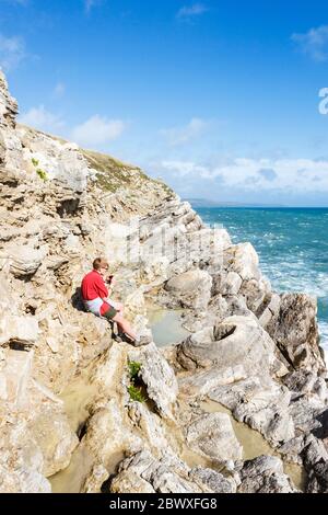 Une jeune fille de quinze ans dans une chemise rouge, assise sur des rochers et prenant des photos dans la forêt fossile sur la côte près de Lulworth, Dorset, Royaume-Uni Banque D'Images