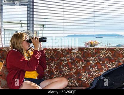 Une jeune fille de 14 ans qui regarde à travers des jumelles vers la mer dans une caravane à Dorset, au Royaume-Uni Banque D'Images