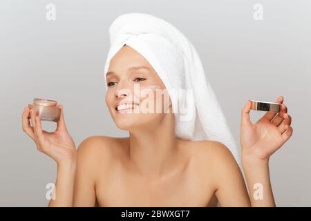 Portrait de jeune femme joyeuse avec serviette blanche sur la tête sans maquillage tenant la crème du visage dans la main regardant avec joie avec la crème sur la joue sur gris Banque D'Images