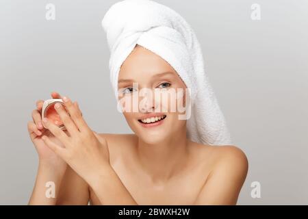 Portrait de jeune femme gaie avec serviette blanche sur la tête sans maquillage application visage crème heureusement regardant dans l'appareil photo sur fond gris isolé Banque D'Images