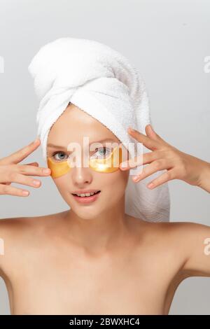Jeune femme attrayante avec serviette blanche sur la tête sans maquillage avec taches jaunes sous les yeux regardant rêveusement dans l'appareil photo sur fond gris isolé Banque D'Images