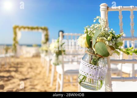 Cérémonie de mariage luxueuse sur l'océan, plage par une journée ensoleillée. Chaises blanches décorées avec un beau bouquet de fleurs dans un pot qui les y est accroché. Banque D'Images