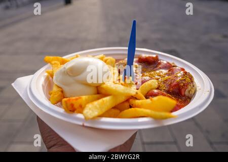 Main tenir Currywurst, saucisses de porc grillées sur le dessus avec poudre de curry, servi avec sauce au ketchup au curry, mayonnaise et pommes de terre frites sur une assiette blanche. Banque D'Images