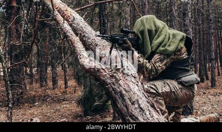 Photo d'un sniper de forêt entièrement équipé à camouflage avec carabine assise et visée près de l'arbre dans les bois. Banque D'Images