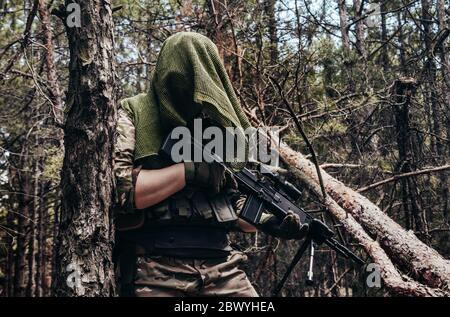 Photo d'un sniper de forêt entièrement équipé à motif camouflage avec carabine debout dans les bois de gros plan. Banque D'Images