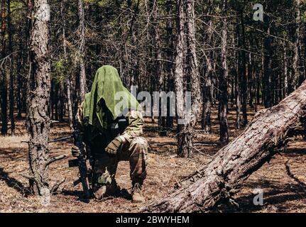 Photo d'un sniper de forêt entièrement équipé à motif camouflage avec fusil et foulard tactique en filet assis dans les bois. Banque D'Images