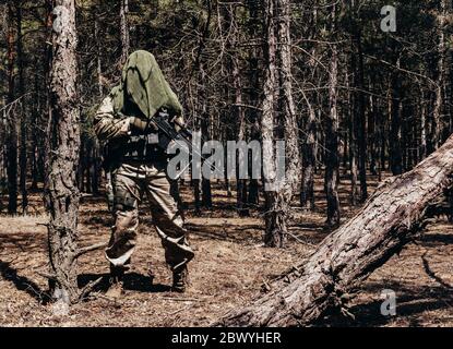 Photo d'un sniper de forêt entièrement équipé à motif camouflage avec carabine et foulard tactique à filet debout dans les bois. Banque D'Images