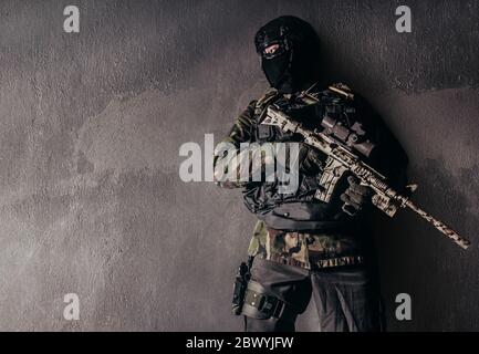 Photo d'un soldat entièrement équipé en uniforme de camouflage debout et appuyé sur un fond de mur en béton. Banque D'Images