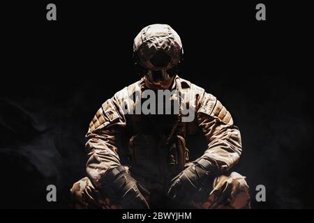 Photo d'un soldat du désert entièrement équipé dans une armure camouflée, un casque et des gants assis sur un fond sombre et brumeux. Banque D'Images