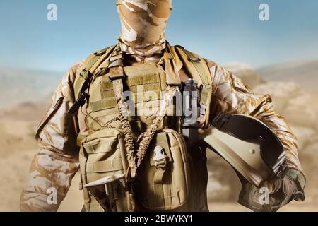 Photo d'un soldat entièrement équipé en uniforme, armure debout dans le désert et tenant un casque avec des lunettes. Banque D'Images