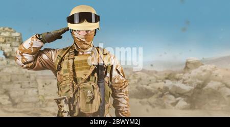 Photo d'un soldat entièrement équipé en uniforme, armure, casque et lunettes debout en salant sur fond de champ de bataille dans le désert. Banque D'Images