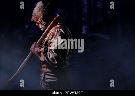 Photo d'un meurtrier sanglant dans une veste de capot marron tenant une hache dans un fond de bois sombre, vue de profil. Banque D'Images