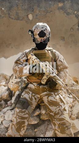 Photo d'un soldat du désert entièrement équipé avec un fusil assis dans une tranchée en pierre avec une armure tactique et un fond de mur endommagé fissuré. Banque D'Images