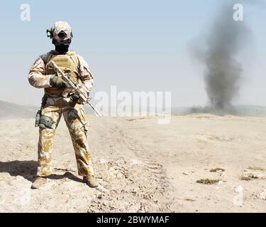 Photo d'un militaire entièrement équipé debout avec une carabine automatique de fond de désert de guerre avec explosion et fumée sur fond. Banque D'Images