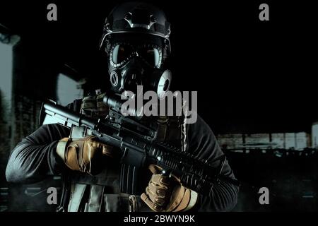 Photo d'un soldat entièrement équipé en gilet tactique d'armure noire, masque à gaz, fusil automatique, gants et casque sur fond sombre ruiné. Banque D'Images