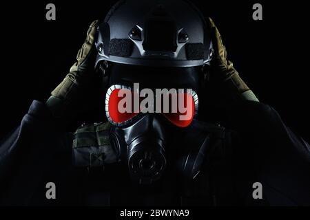 Photo d'un soldat entièrement équipé en gilet tactique d'armure noire, masque à gaz avec yeux rouges, gants et mise sur casque debout sur fond noir clos Banque D'Images