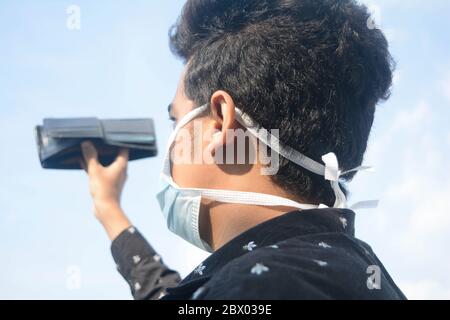 Jeune homme portant un masque facial de protection et montrant son portefeuille vide sur fond de nuages et de ciel. Banque D'Images