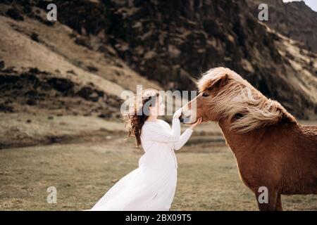 Une mariée vêque d'une robe blanche se fond le nez d'un cheval. Les cheveux et la manie se développent dans le vent. Destination Islande mariage séance photo avec des chevaux islandais Banque D'Images