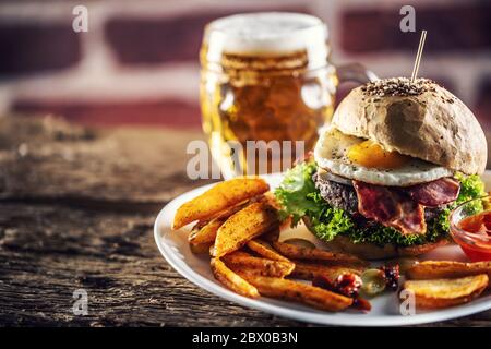 Hamburger avec un pain au sésame, un œuf et du bacon frits, une salade et des quartiers de pommes de terre avec une bière pression à l'arrière-plan Banque D'Images