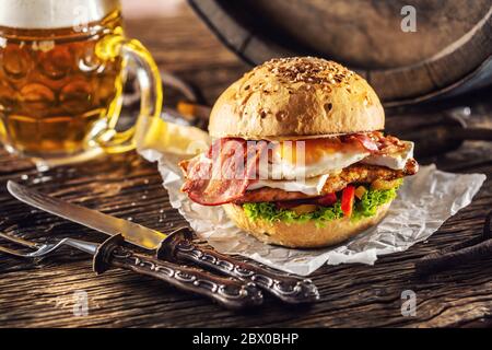 Hamburger au poulet, œuf et bacon frits, salade, poivrons avec un couteau rustique et une fourchette sur le côté avec une bière pression Banque D'Images