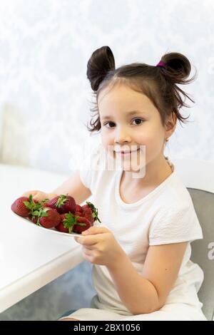 une belle petite fille en robe blanche tient une assiette avec les premières fraises. printemps. mai. pandémie Banque D'Images