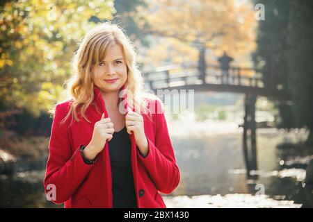 jeune femme attrayante portant un manteau rouge dans le parc de la ville d'automne près du lac. regardant vers l'avant dans la caméra Banque D'Images