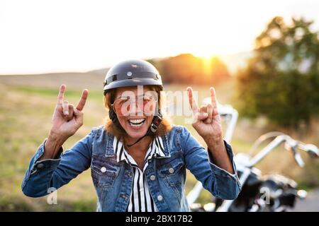 Une femme âgée et gaie voyageant à moto à la campagne au coucher du soleil. Banque D'Images