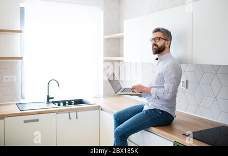 Un homme mature avec ordinateur portable assis dans la cuisine dans une nouvelle maison non meublée. Banque D'Images