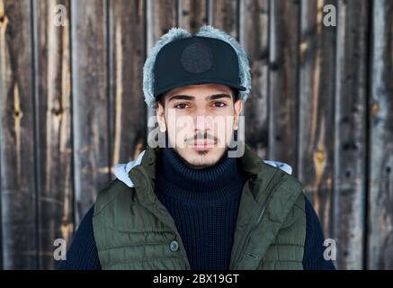 Jeune homme avec un chapeau debout contre un fond en bois à l'extérieur en hiver. Banque D'Images