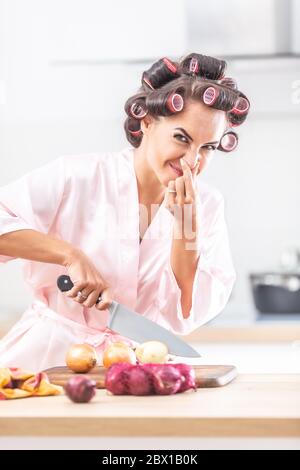 Femme en chemise de nuit et les rouleaux de cheveux tient son nez d'un oignon malodorant qu'elle coupe sur une planche à découper. Banque D'Images
