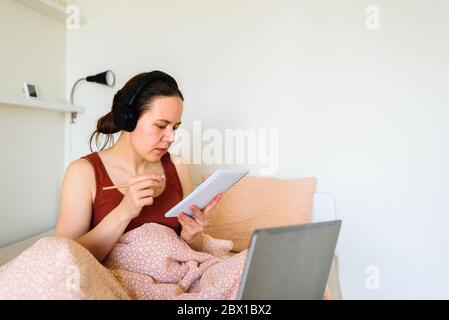 Femme souriante travaillant de la maison dans le lit. Écriture dans le bloc-notes. La vraie vie, le bureau dans la chambre Banque D'Images
