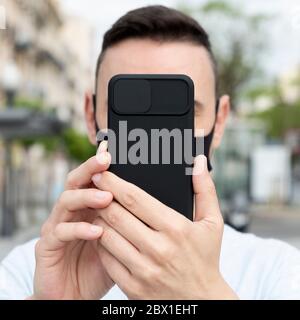 gros plan sur un jeune homme du caucase dans la rue, portant un visage noir prenant un selfie ou faisant un appel vidéo avec son smartphone, placé devant lui Banque D'Images