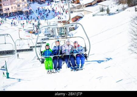 Quatre amis enfants s'assoient sur le télésiège de la montagne avec la piste bondée et la station en dessous Banque D'Images