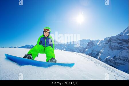 Un petit garçon mignon s'assoit sur la vue du snowboard sur le ciel bleu et les sommets de montagne à l'arrière Banque D'Images