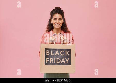 Portrait de jeune femme avec des cheveux bouclés souriant à l'appareil photo tout en tenant l'achat du Vendredi fou sur fond rose Banque D'Images