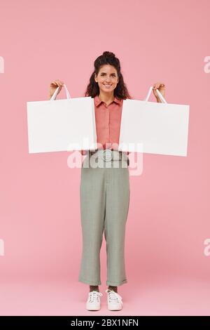Portrait d'une jeune femme aux cheveux bouclés souriant à l'appareil photo et tenant deux sacs d'achats dans ses mains contre le fond rose Banque D'Images