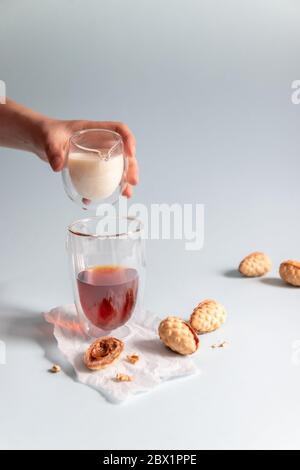 Une main femelle verse du lait dans du thé noir dans une tasse de verre. Biscuits de noyer sur fond bleu, photo verticale Banque D'Images