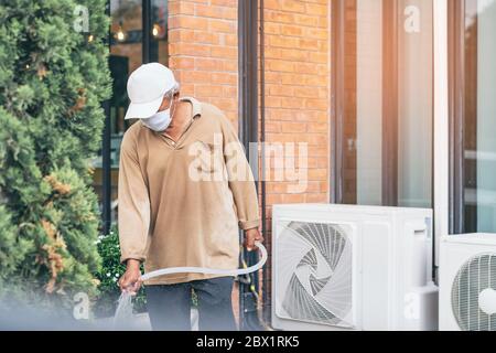 Un homme âgé en quarantaine porte un masque facial pour empêcher la propagation du virus Corona (Covid-19) arroser des plantes pour faire de l'exercice dans le jardin à la maison. Ne Banque D'Images