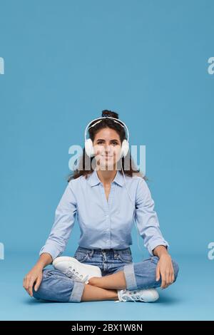 Portrait d'une jeune femme dans un casque assis sur le sol et souriant à l'appareil tout en écoutant de la musique sur fond bleu Banque D'Images
